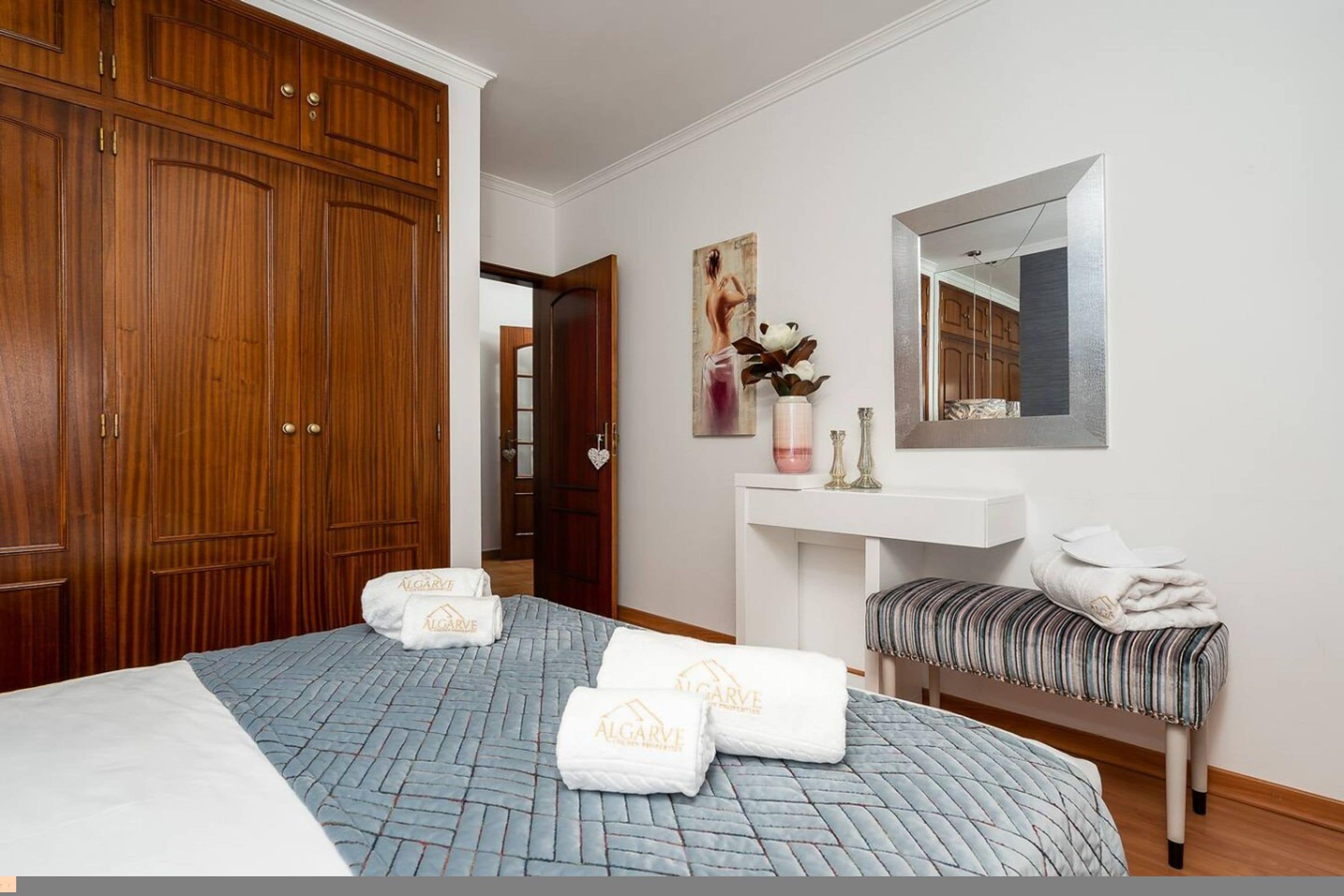 Una camera d'albergo con un'ampia armadiatura in legno, un letto con copriletto blu, un comodino bianco con specchio e una panca con asciugamani.