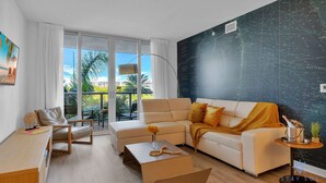 A modern living room with a sofa, armchair, and coffee table. There is a large window with a view of palm trees and buildings. - Spectacular Near Beach - 2 BR Apartment (Hallandale Beach)