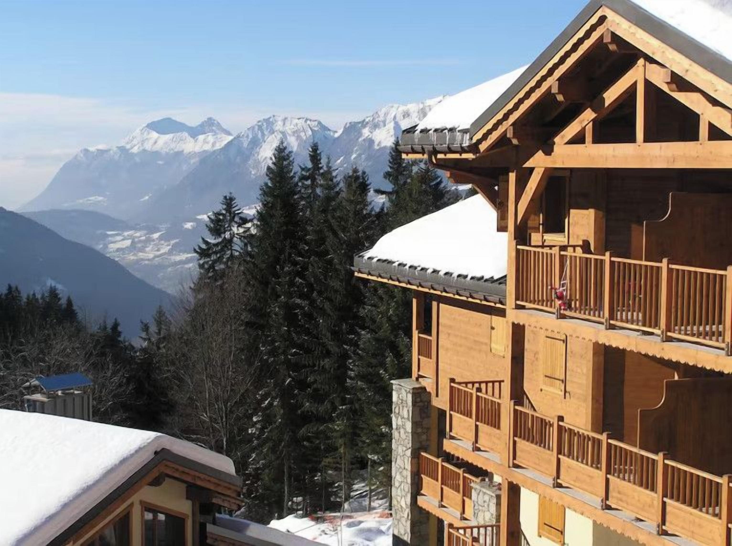 A wooden chalet with a balcony, set against a backdrop of snow-covered mountains.