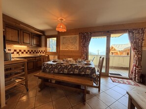 Une cuisine chaleureuse avec des meubles en bois, une table à manger dressée pour quatre personnes et une vue sur les montagnes par la porte ouverte.