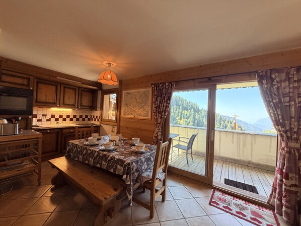 Une cuisine chaleureuse avec des meubles en bois, une table à manger pour quatre personnes et une grande fenêtre donnant sur un paysage montagneux.