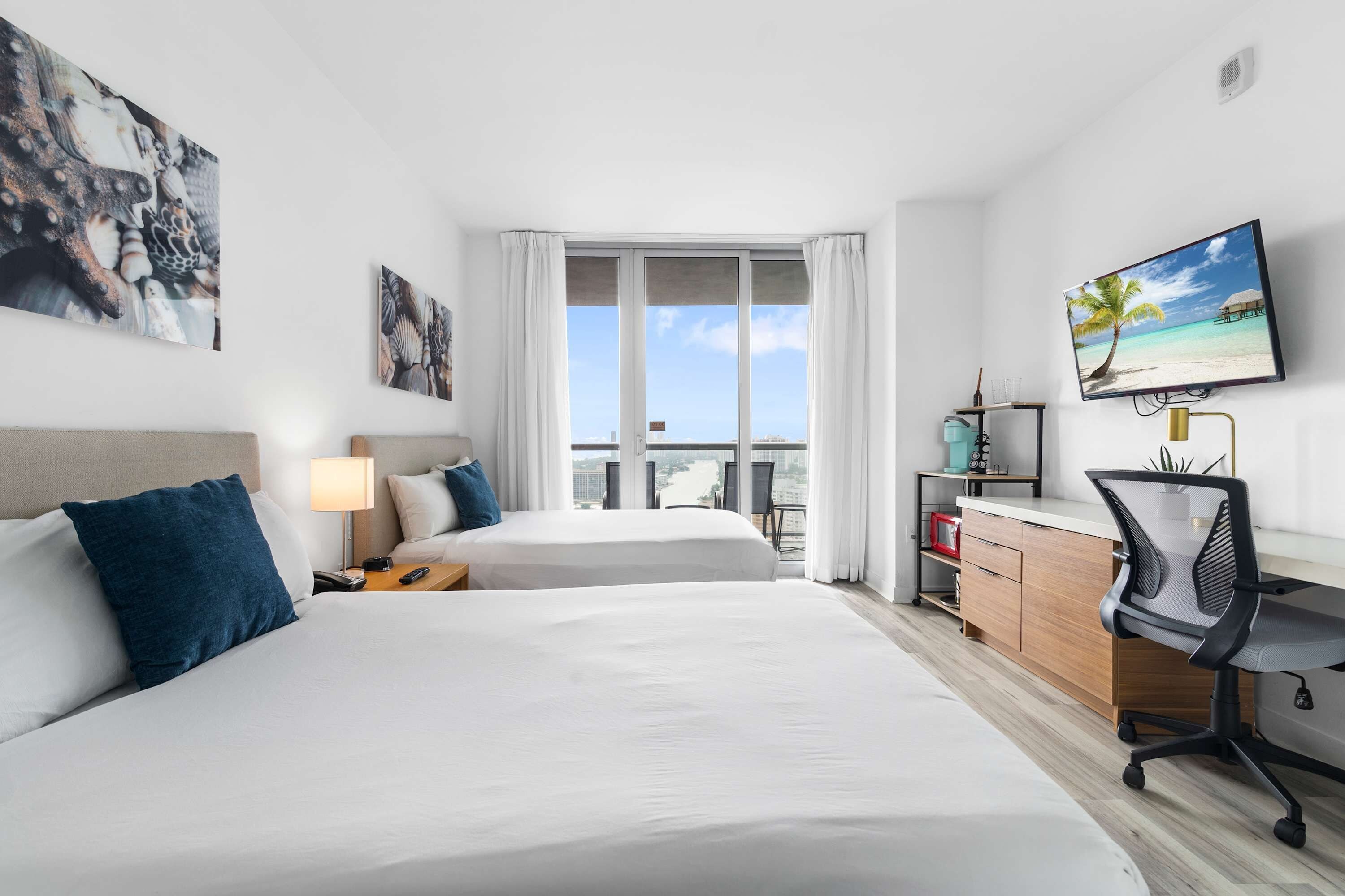 A hotel room with two beds, a desk, a chair, a TV, and a window with a view of the city.