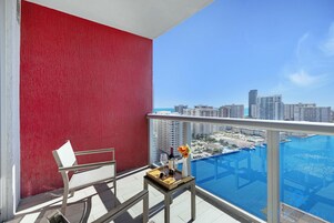 Un balcón con pared roja, una silla blanca, una mesita con una botella y un vaso, y vistas a un horizonte urbano y un puerto deportivo.