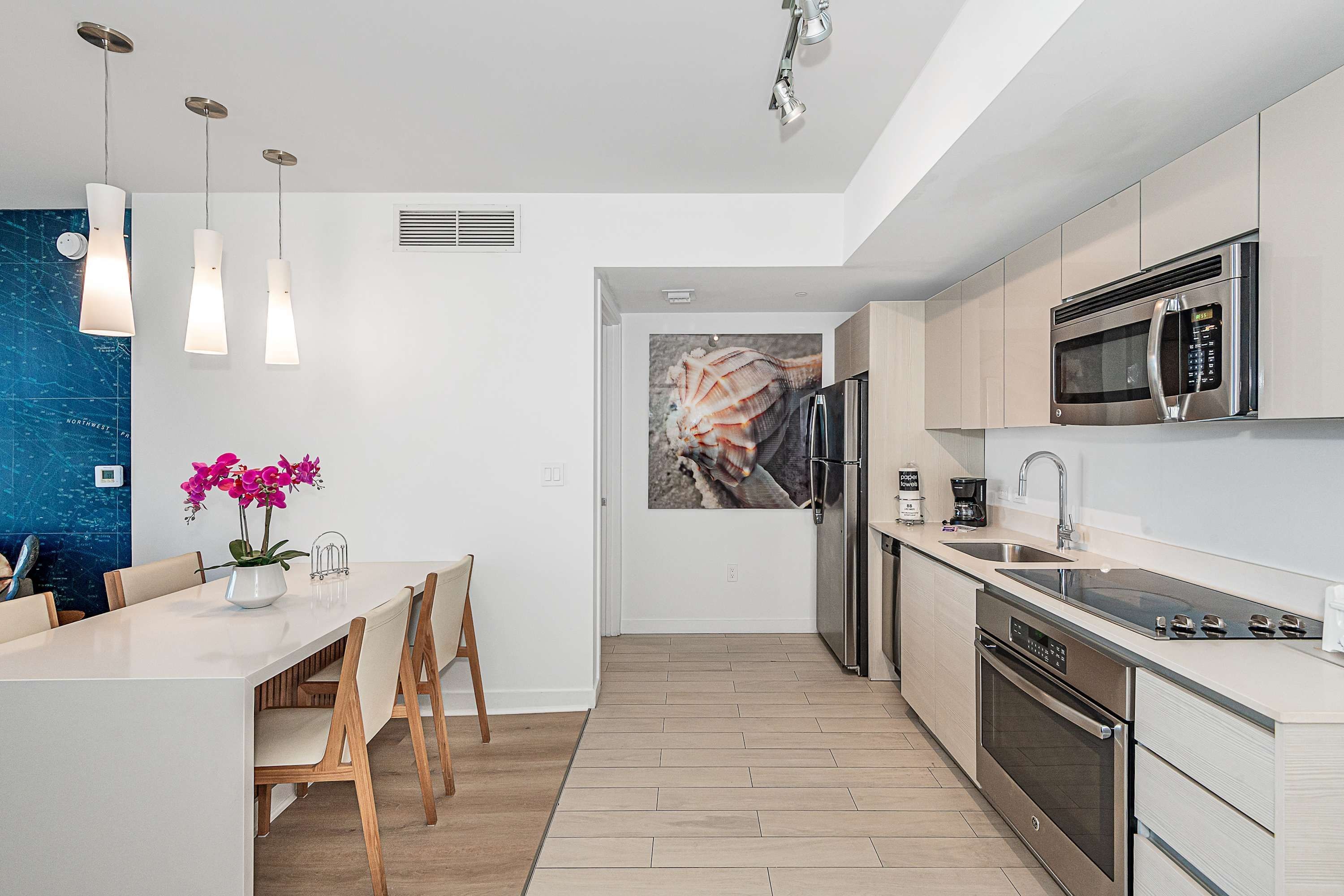 A modern kitchen with a built-in microwave, oven, and sink. There is a dining area with chairs and a table, and a decorative wall with a seashell painting.