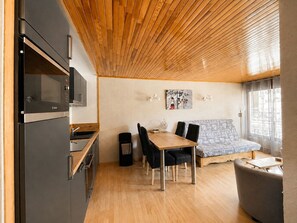 A modern kitchen with a built-in oven and microwave, a dining area with a wooden table and chairs, and a sofa with a patterned cover. - Le Cygne - 6-guest balcony apartment (Les Deux Alpes)