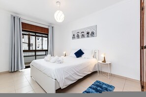 Ein Hotelzimmer mit Bett, Nachttisch, blauem Teppich, Fenster mit Vorhängen und Deckenlampe.