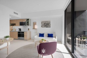 A modern living space with a kitchen, a white sofa with blue cushions, a purple chair, and a dining area with a table and chairs. - 1BR Apartment - Pool Gym (Miami)