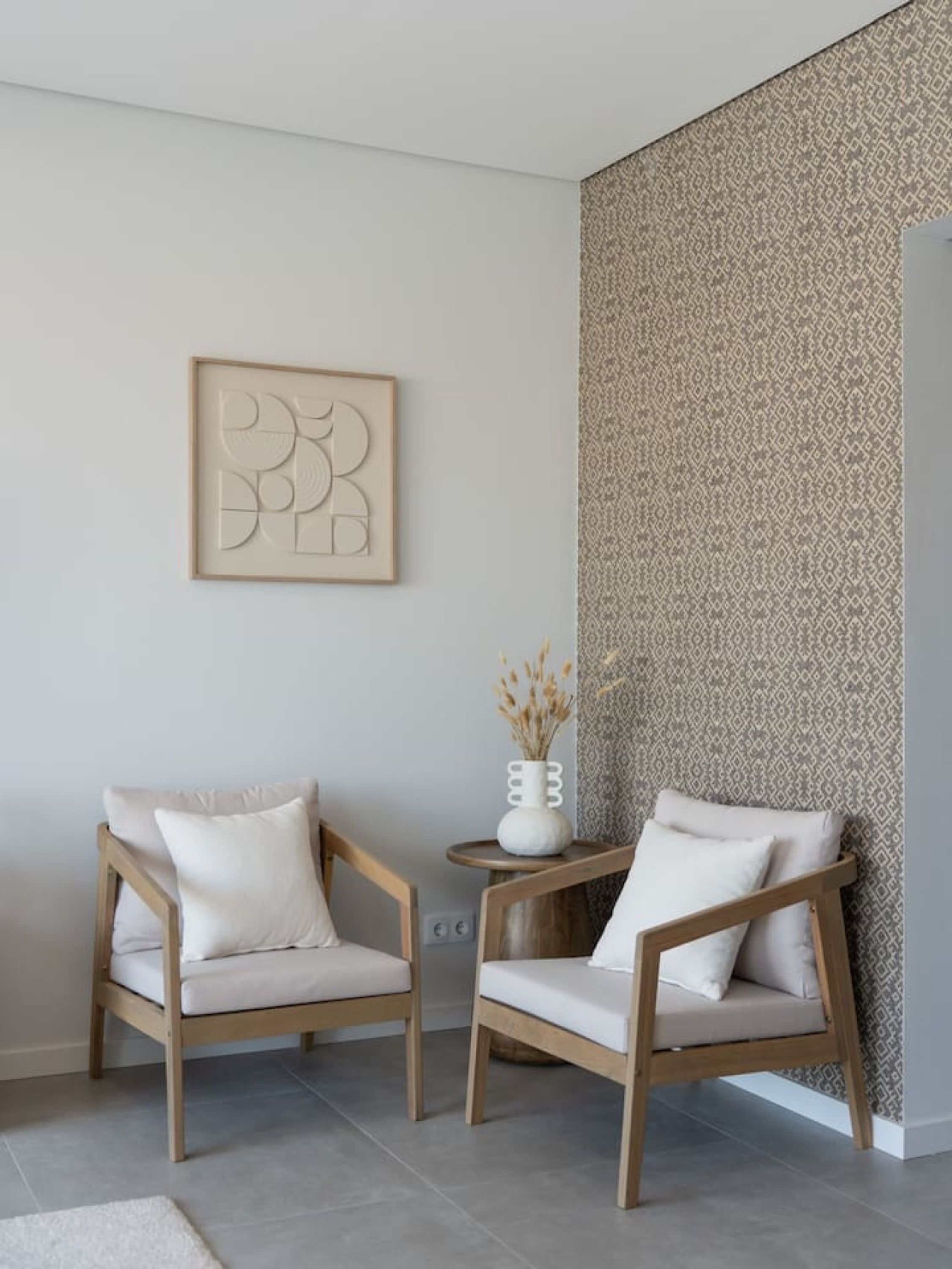 A room with two wooden chairs with cushions, a small round table with a vase, and a patterned wallpaper on the wall.