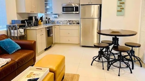 A modern kitchen with stainless steel appliances, a dining area with a round table and stools, and a living space with a leather sofa and ottoman. - Beachside Comfort 2BR and 2BA With Outdoor Dining (Hollywood)
