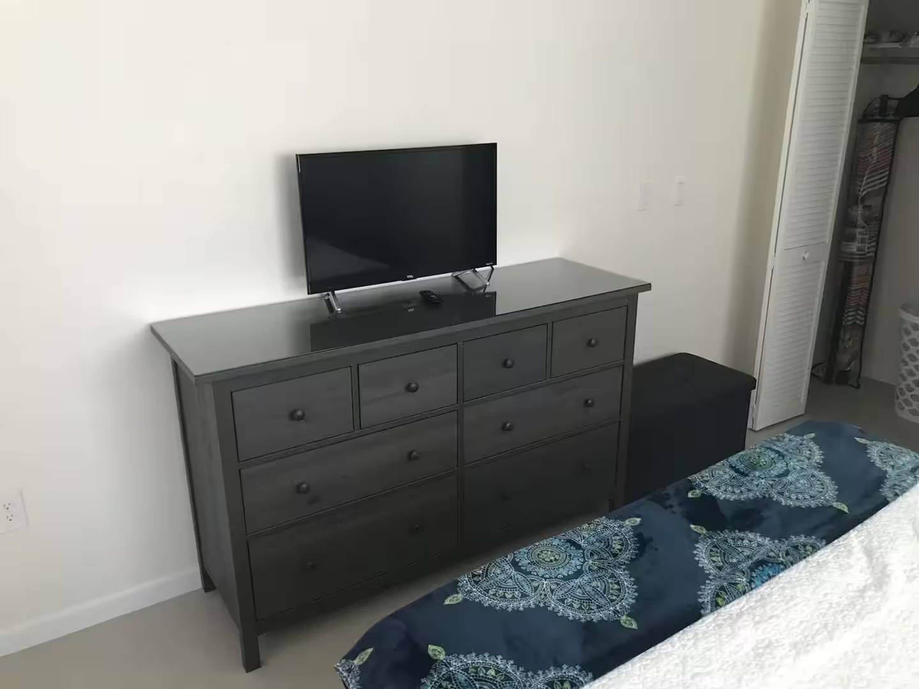 A bedroom with a dark dresser, a flat-screen TV, a patterned bedspread, and a white door in the background.