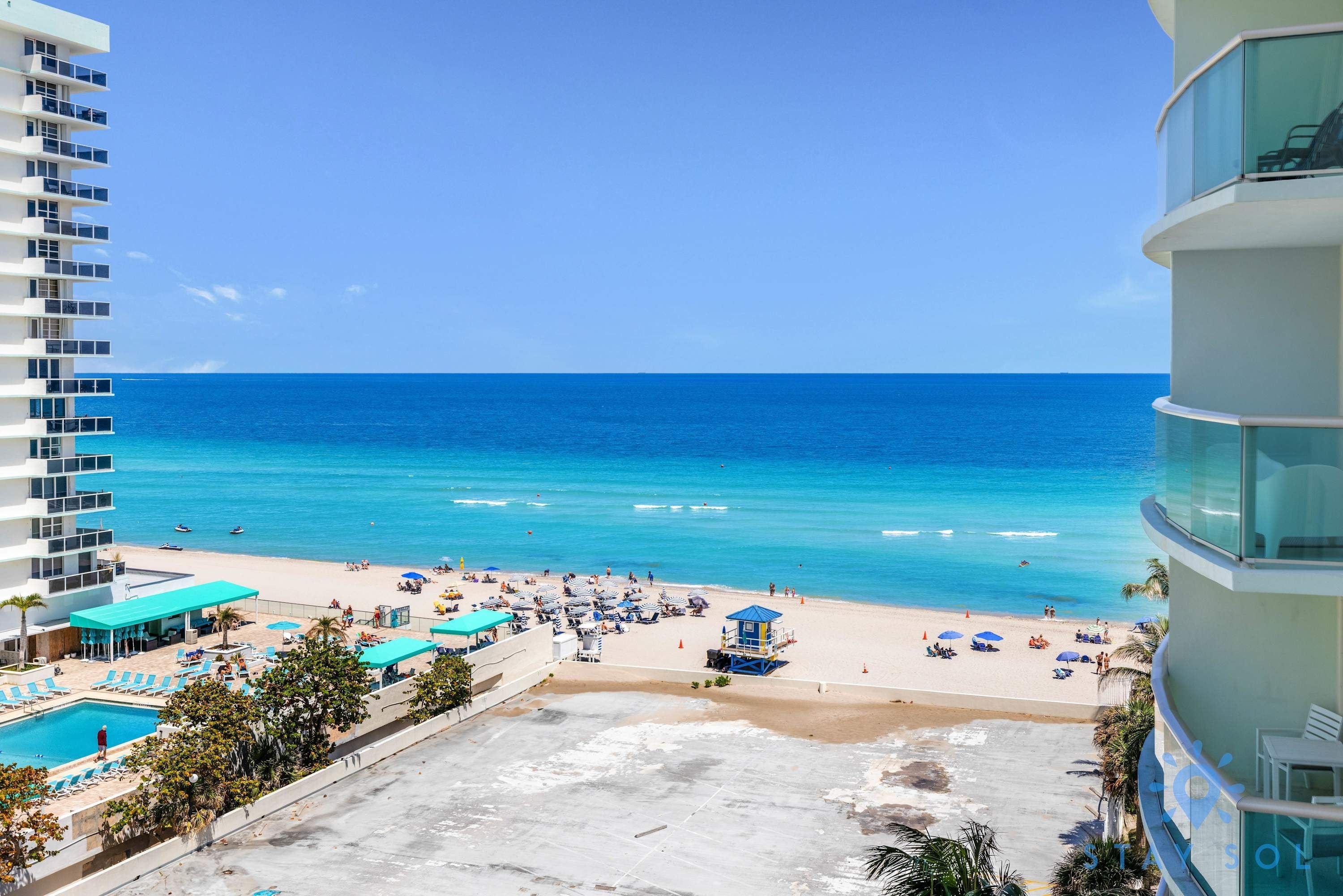 A beachfront hotel with a pool, beach umbrellas, and a lifeguard tower.