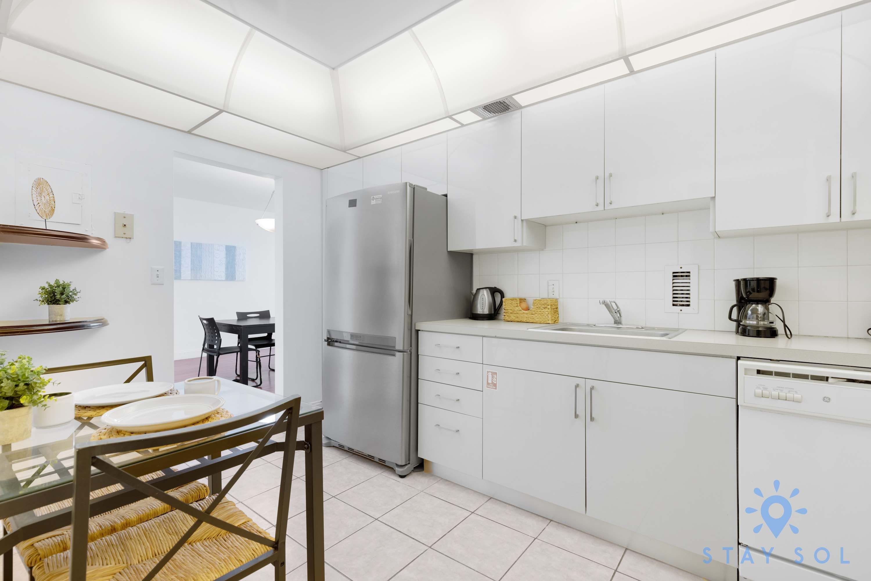 A modern kitchen with white cabinets, a stainless steel refrigerator, and a dining area with a table and chairs.
