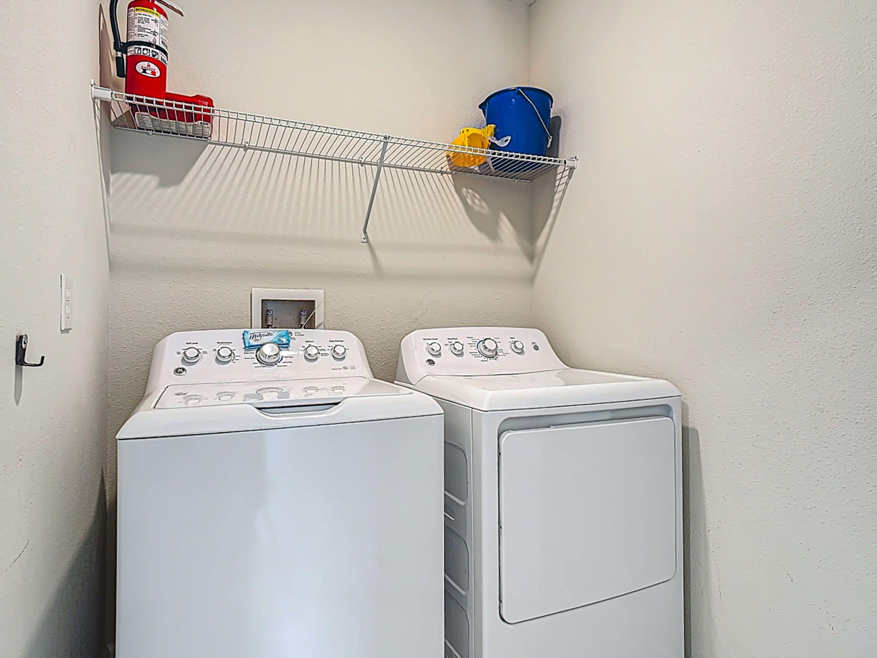 A laundry room with two front-loading washing machines, a fire extinguisher, and a metal shelf with a blue bucket and yellow cloth.