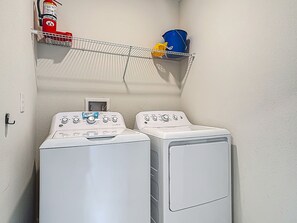 A laundry room with two front-loading washing machines, a fire extinguisher, and a metal shelf with a blue bucket and yellow cloth. - Stylish 3-bedroom Townhouse in Champions Gate Sleeps 12 Resort Amenities (Almont)
