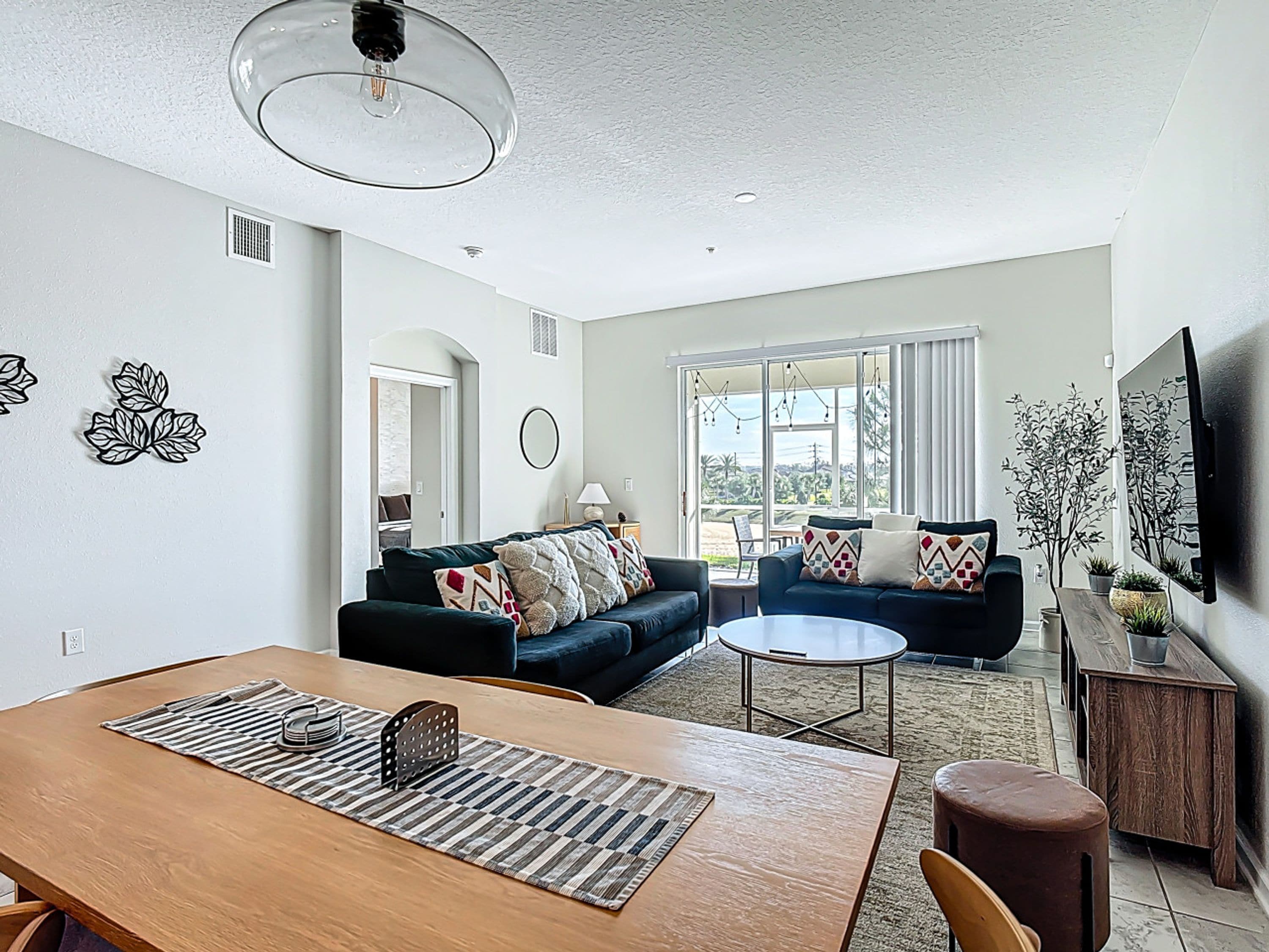 A modern living room with a dining area, large windows, and a flat-screen TV mounted on the wall.