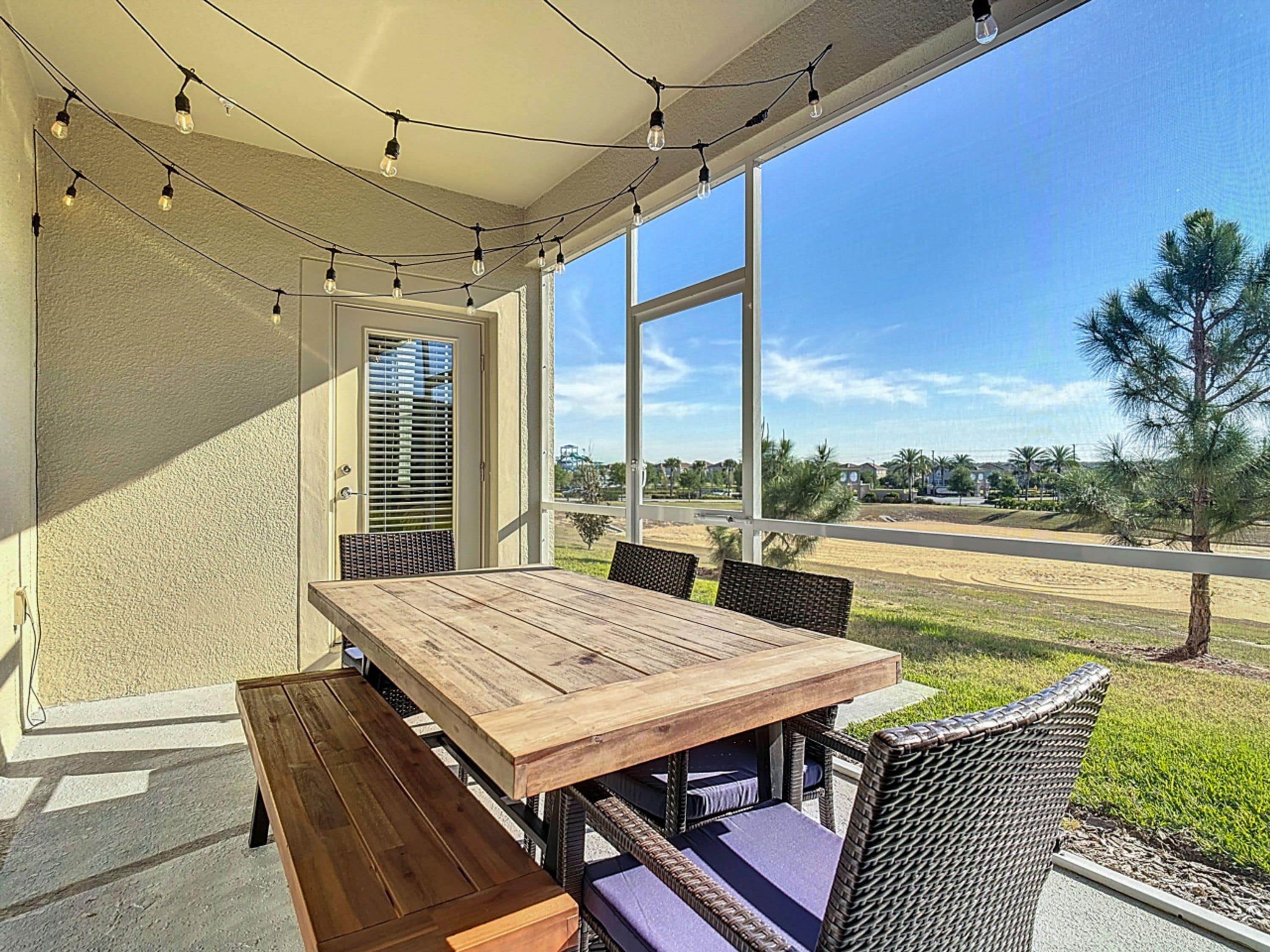 Een overdekt terras met een houten tafel en stoelen, sfeerverlichting en uitzicht op een woonwijk.