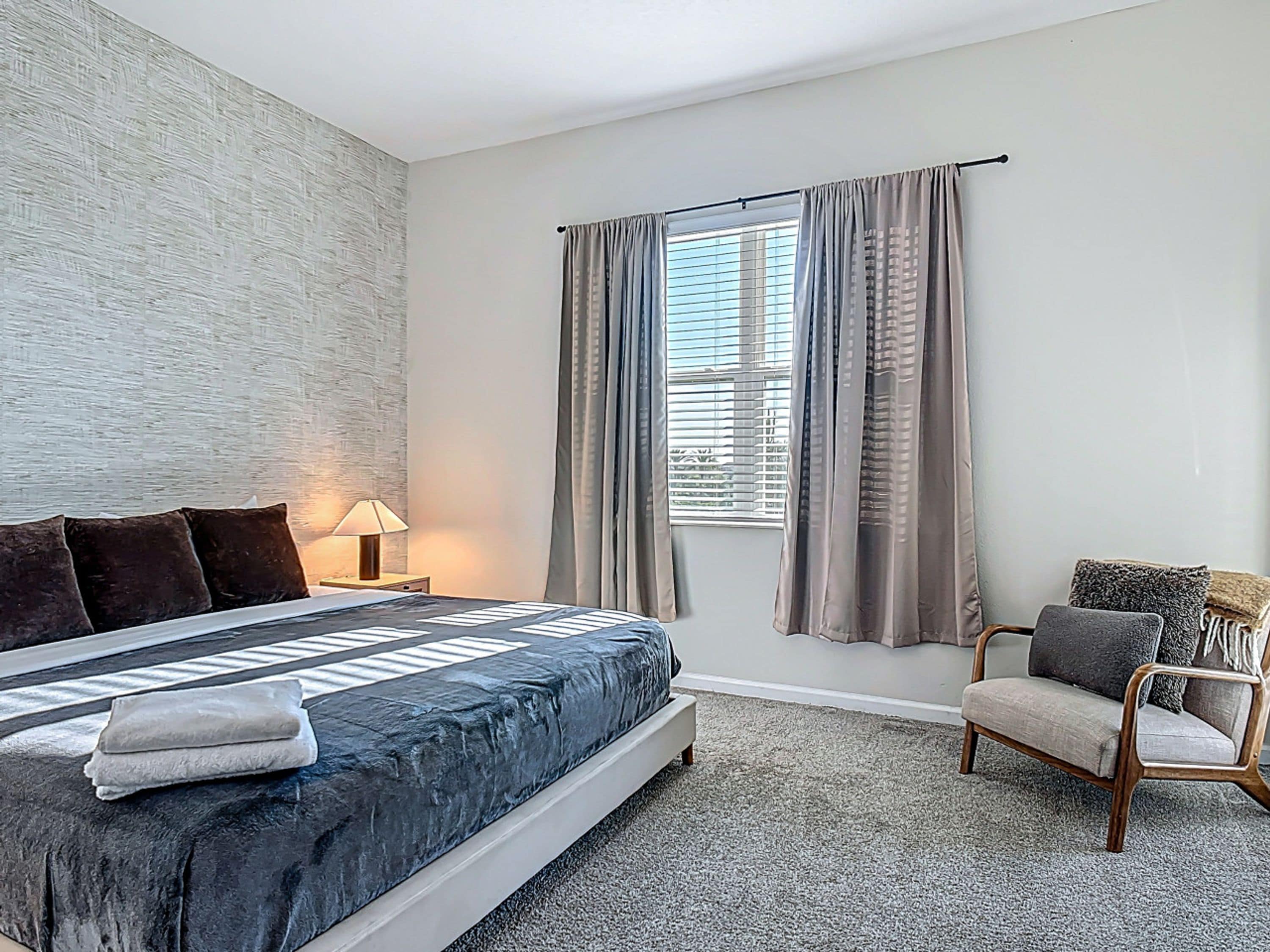 A bedroom with a bed, a chair, a nightstand, a lamp, and a window with curtains.