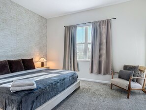 A bedroom with a bed, a chair, a nightstand, a lamp, and a window with curtains. - Stylish 3-bedroom Townhouse in Champions Gate Sleeps 12 Resort Amenities (Almont)
