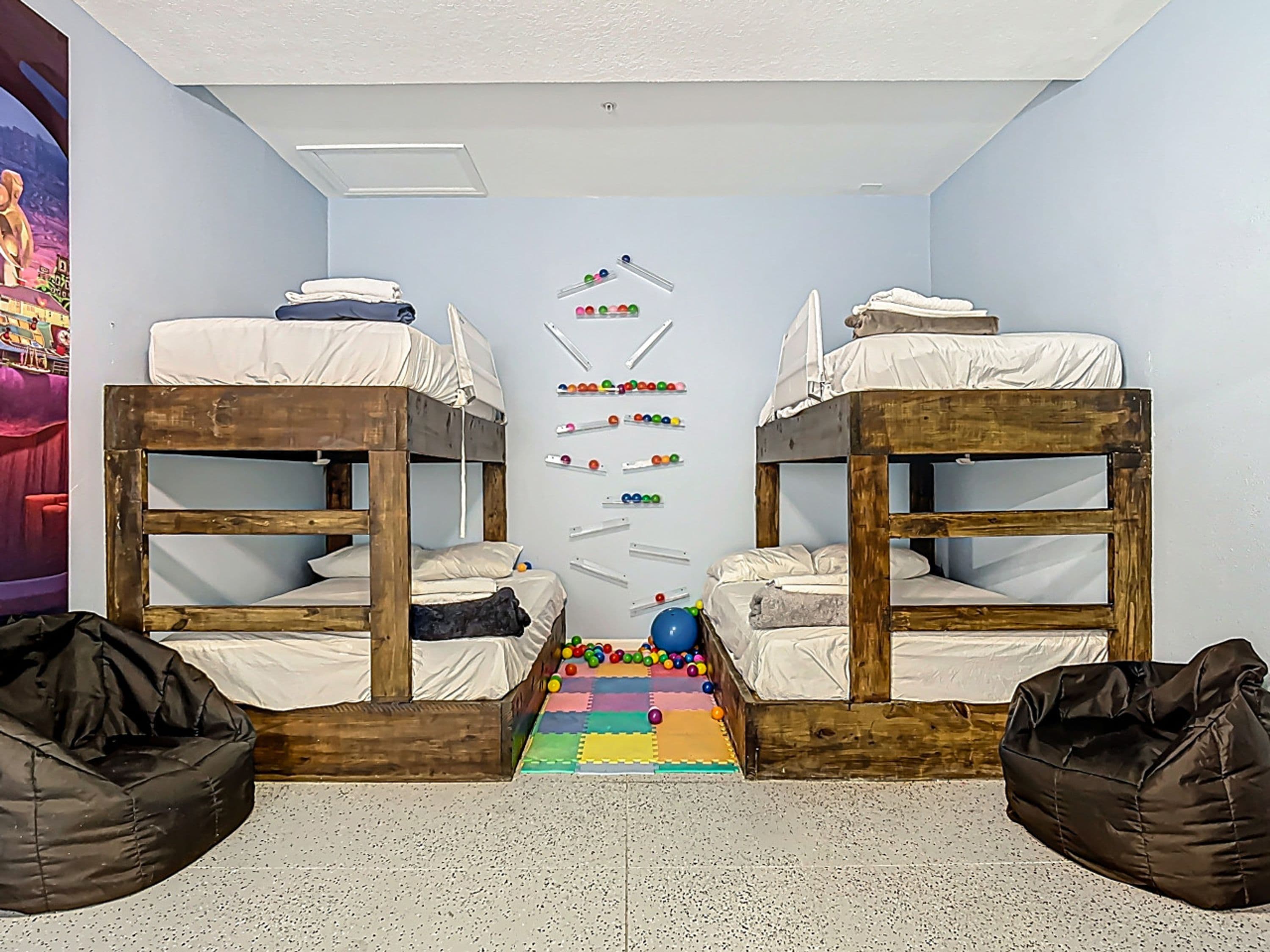 A room with two bunk beds, a colorful play mat, and a wall with a ball rack.