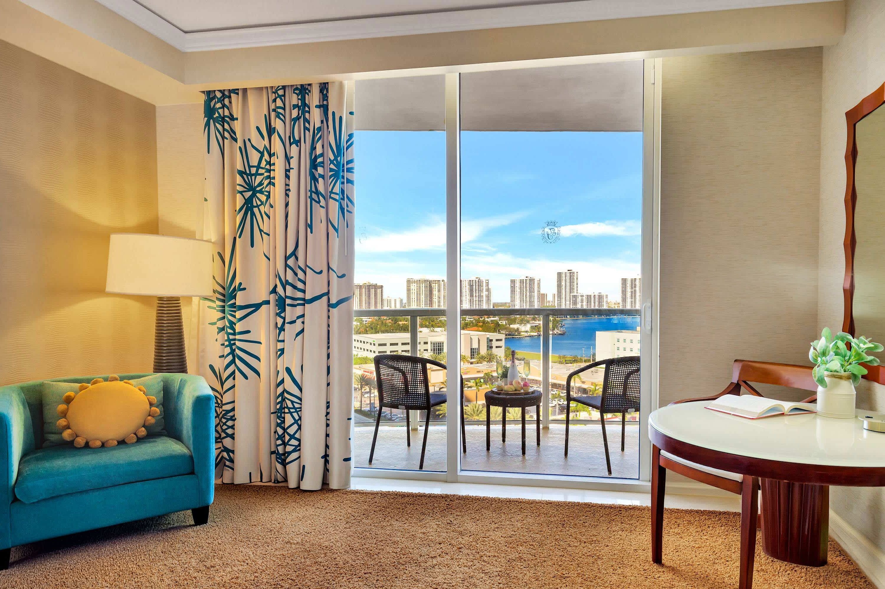Habitación de hotel con sofá azul, mesa de comedor con sillas, ventana grande con vistas a la ciudad y al agua, y cortina con estampado de hojas.
