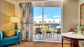 Habitación de hotel con sofá azul, mesa de comedor con sillas, ventana grande con vistas a la ciudad y al agua, y cortina con estampado de hojas.