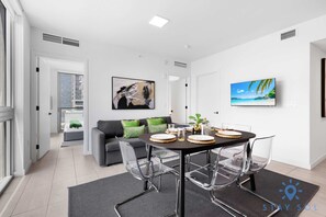 A modern dining area with a round table, clear chairs, and a view of the city through a large window. - Beautiful 2BR Apartment With gym and Pool (Miami)