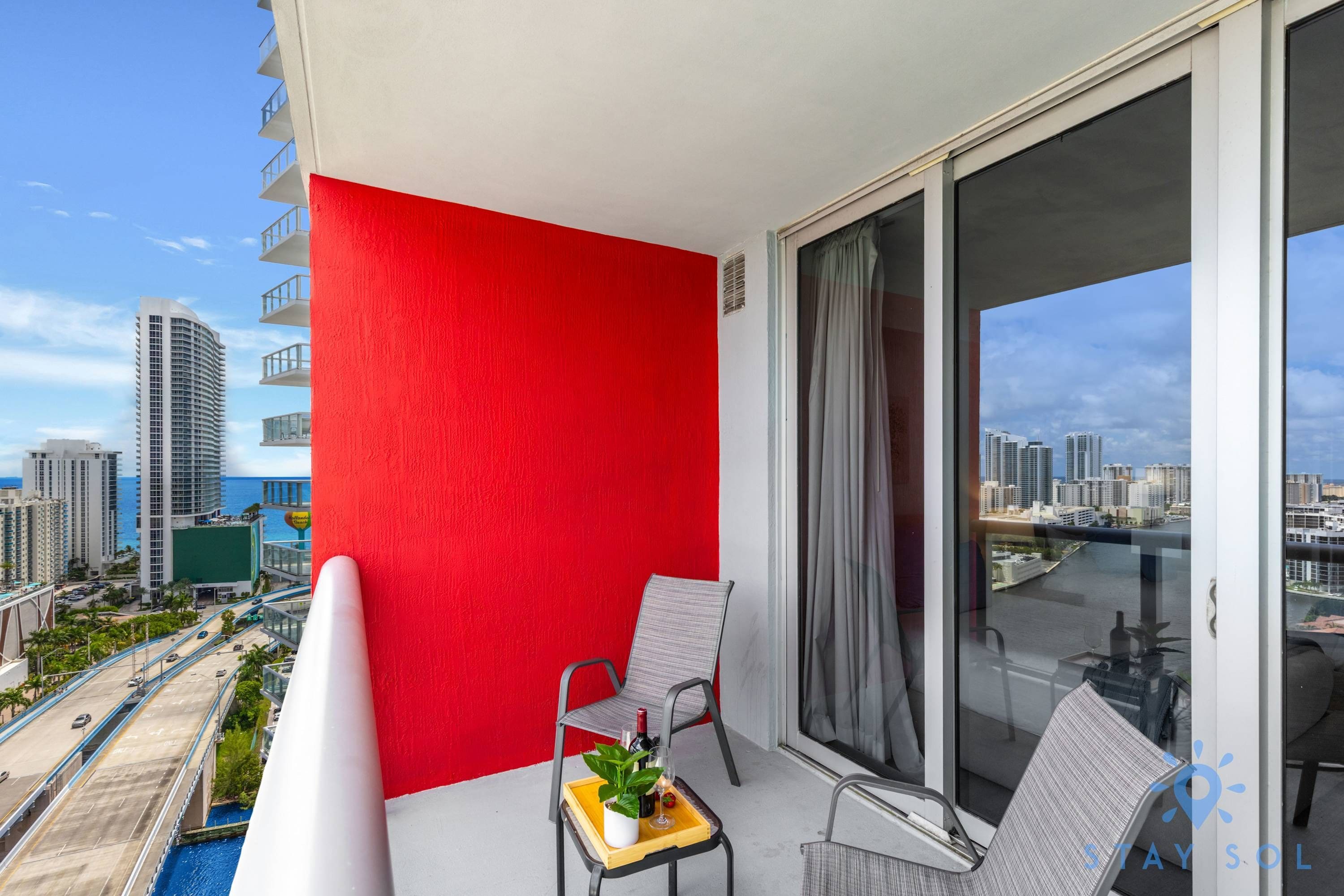 Un balcone con un muro rosso, due sedie, un tavolino con una pianta e vista sulla città.