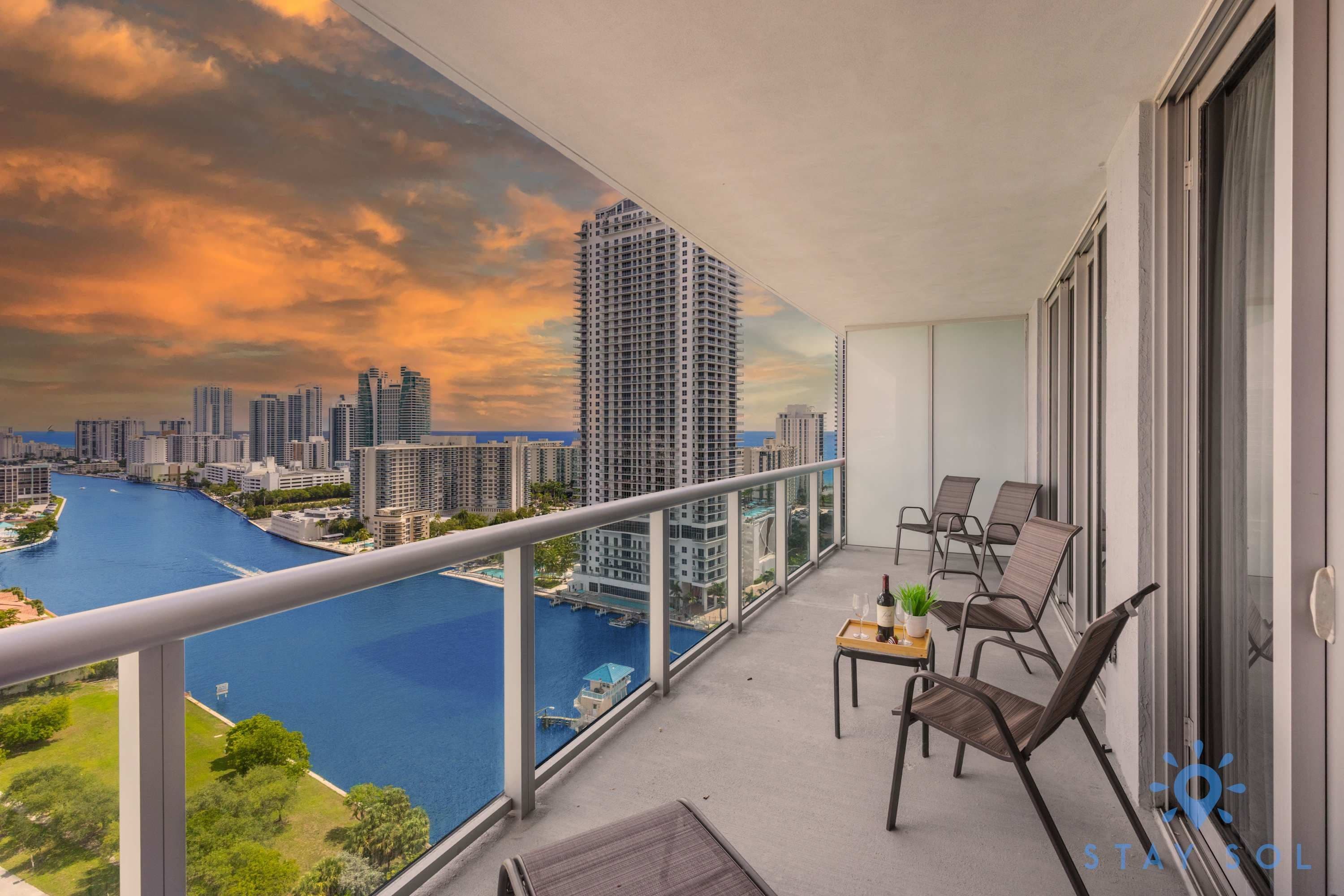 A balcony with a view of a city skyline and a river. There are chairs and a small table with a bottle and glass.