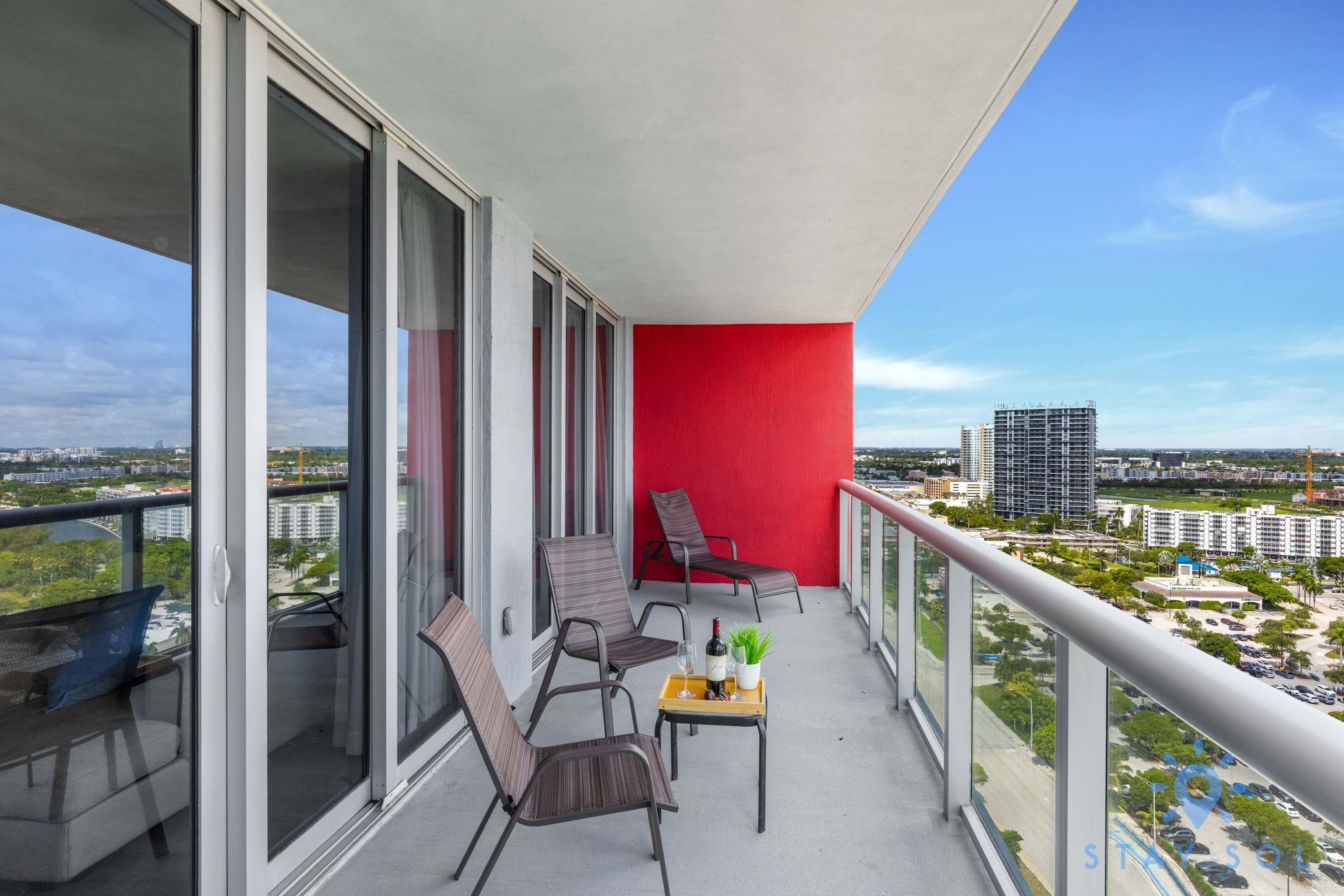 A balcony with a red wall, two chairs, a small table with a bottle and glasses, and a view of the cityscape.