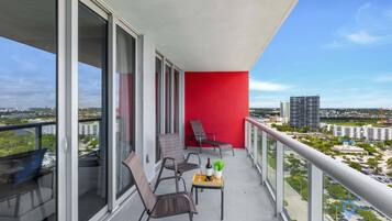 A balcony with a red wall, two chairs, a small table with a bottle and glasses, and a view of the cityscape.
