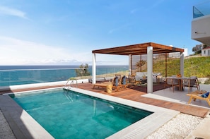 A swimming pool with a wooden deck and lounge chairs, overlooking a body of water with mountains in the distance. - Villa Lara Seafront in Polis (Pachyammos)
