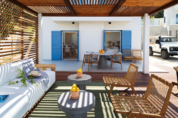 A covered outdoor seating area with a sofa, chairs, and a table, a white vehicle parked nearby, and a building with blue shutters in the background. - Villa Lara Seafront in Polis (Pachyammos)
