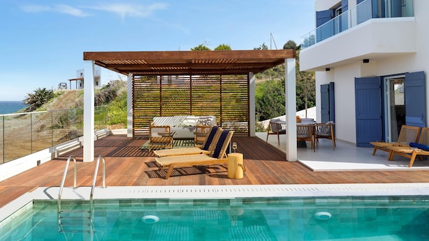 A modern outdoor pool area with a wooden deck, lounge chairs, and a covered seating area. - Villa Lara Seafront in Polis (Pachyammos)