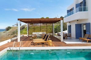 A modern outdoor pool area with a wooden deck, lounge chairs, and a covered seating area. - Villa Lara Seafront in Polis (Pachyammos)
