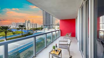A balcony with a red wall, a table with a bottle and glass, and a view of a cityscape with buildings and a blue body of water.