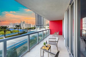 Un balcón con una pared roja, una mesa con una botella y un vaso, y vistas de un paisaje urbano con edificios y una masa de agua azul.