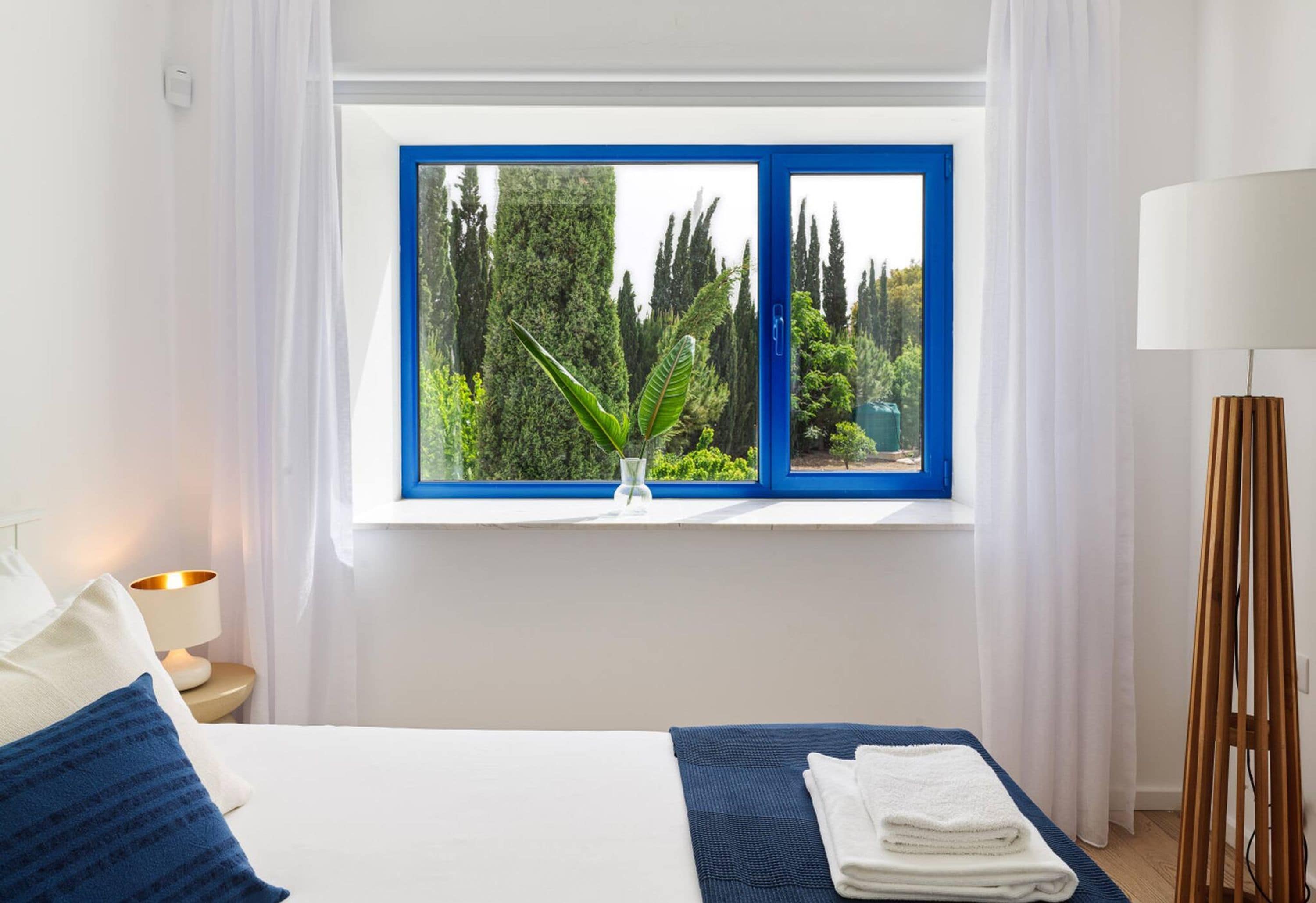 A bedroom with a bed, a nightstand, a lamp, a window with a view of greenery, and a bedside table.