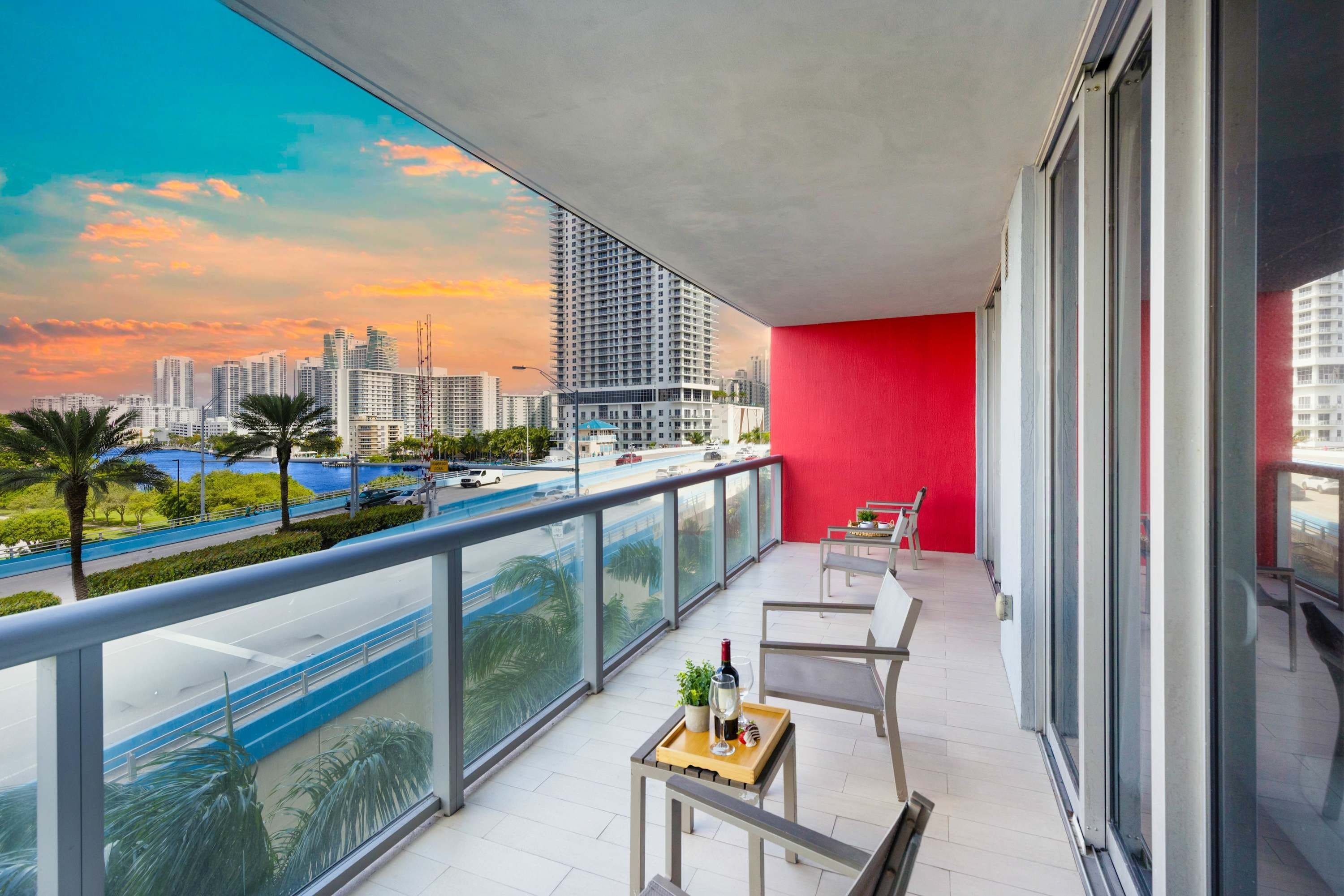 Uma varanda com parede vermelha, mesa com garrafa e copo, e vista para um horizonte urbano com edifícios e um corpo d'água azul.