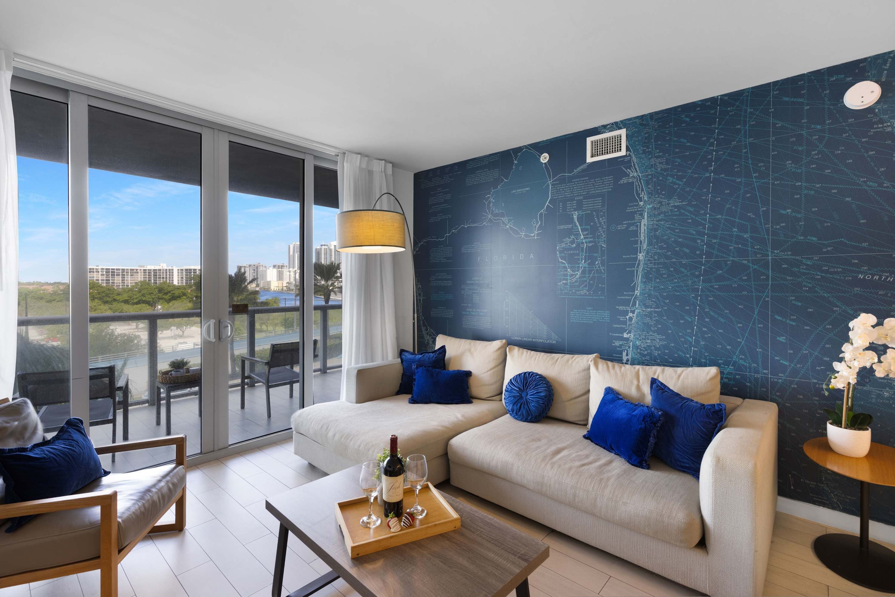 Una sala moderna con un ventanal grande, un mural de mapa, un sofá con cojines azules y una mesa de comedor con una botella y vasos.