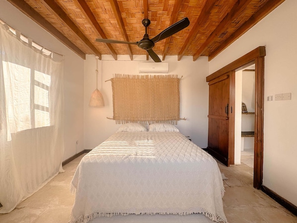 Une chambre à coucher avec un lit, un ventilateur de plafond, une décoration murale en fibres tressées et une porte donnant sur une autre pièce.