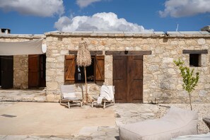 A stone building with a large wooden door, two lounge chairs with cushions, and a small tree in the foreground. - Soumaki Countryside Villa (Pano Akourdalia)
