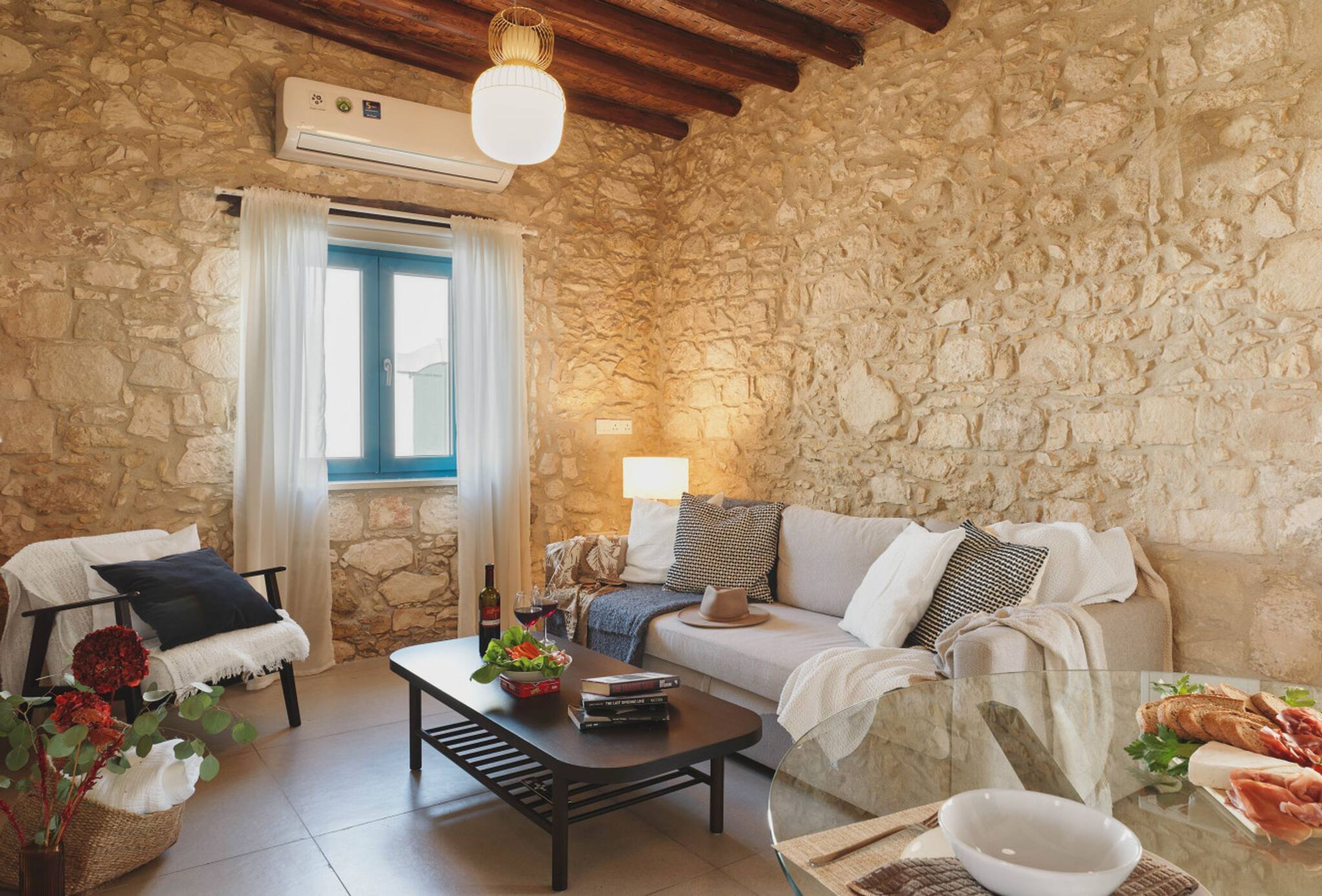 Una habitación acogedora con paredes de piedra, techo de madera, un sofá, una mesa de centro y una zona de comedor con pan y queso.