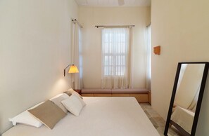 Un dormitorio con cama, lámpara de noche, ventana con cortinas, banco y espejo.
