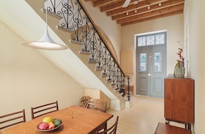 Un comedor con una mesa de madera, sillas y un frutero. Una escalera con barandillas de metal decorativas conduce a una puerta con paneles de vidrio.