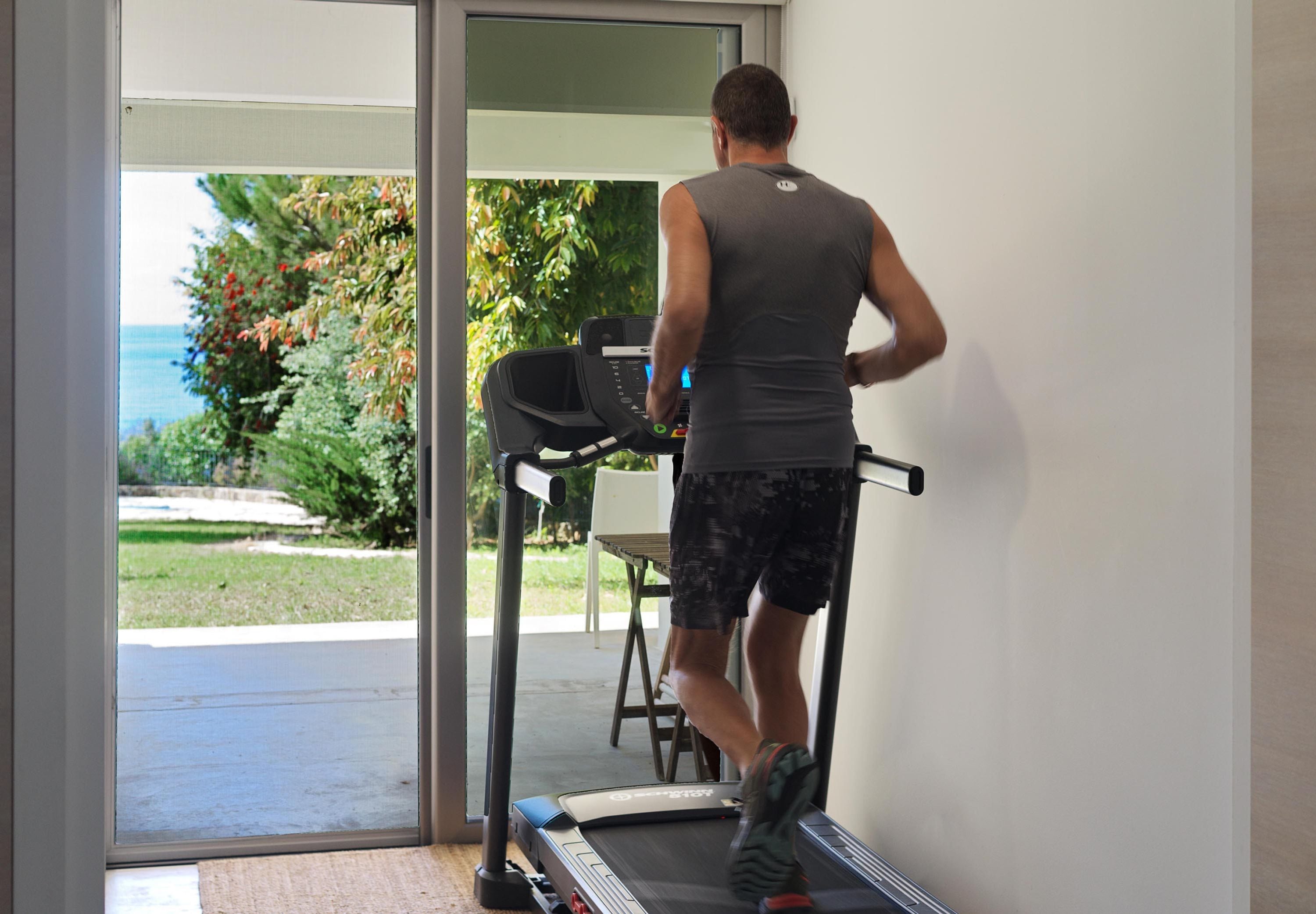 Una persona corre en una cinta de correr dentro de una habitación con vistas a un jardín y un cuerpo de agua.