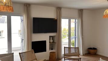 Una sala de estar con un sofá, dos sillones de mimbre, una mesa de centro, un televisor de pantalla plana montado en la pared y un ventanal con cortinas.