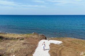 A coastal landscape with a clear sky, a rocky shoreline, and a person walking on a gravel path. - Villa Paradise in Polis (Pachyammos)