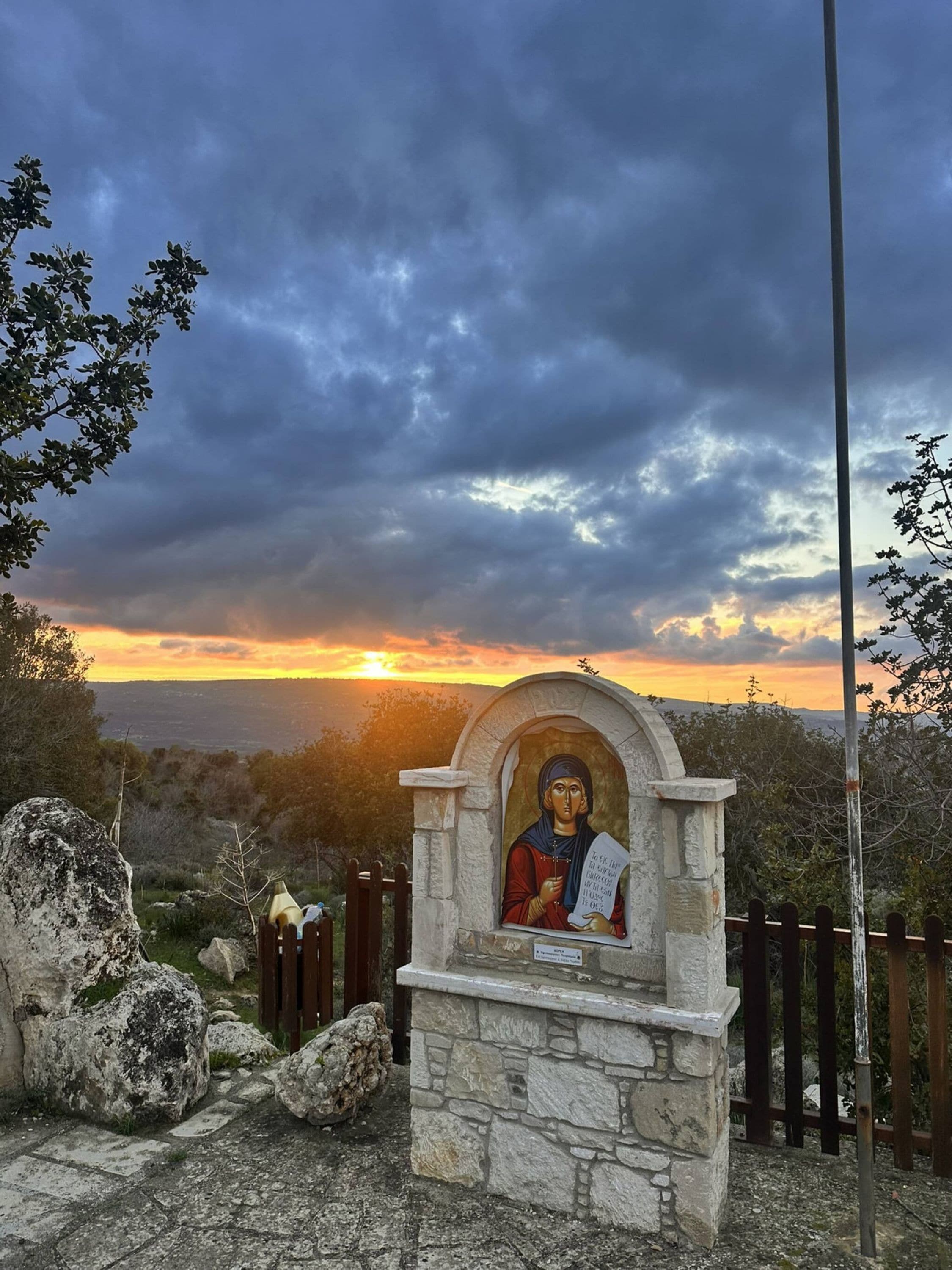 Una estructura de piedra con un icono religioso, rodeada por una valla de madera y rocas, con vistas a un paisaje con un atardecer de fondo.