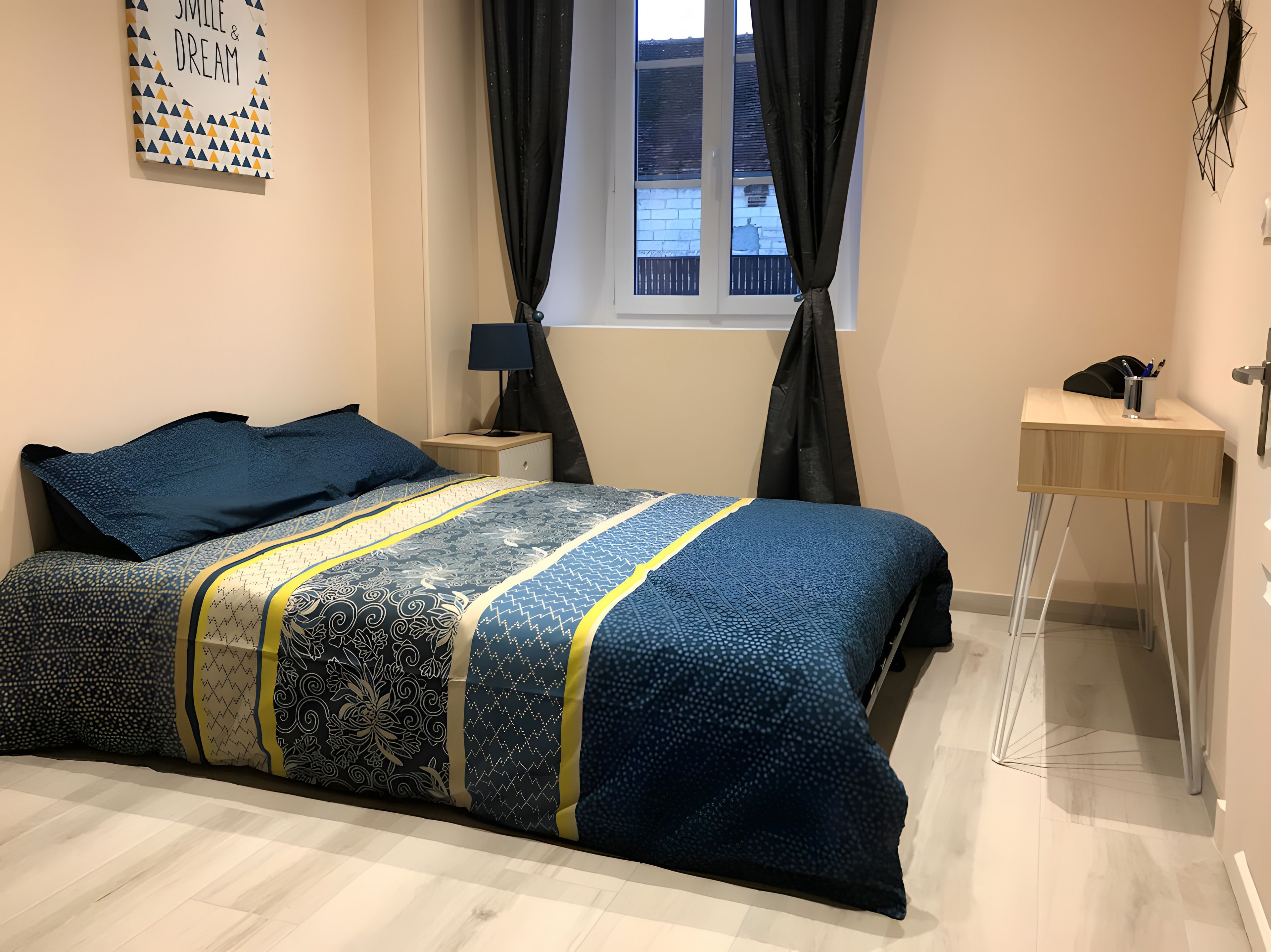 A bedroom with a bed, a nightstand, a desk, a window with curtains, and a wall decoration.