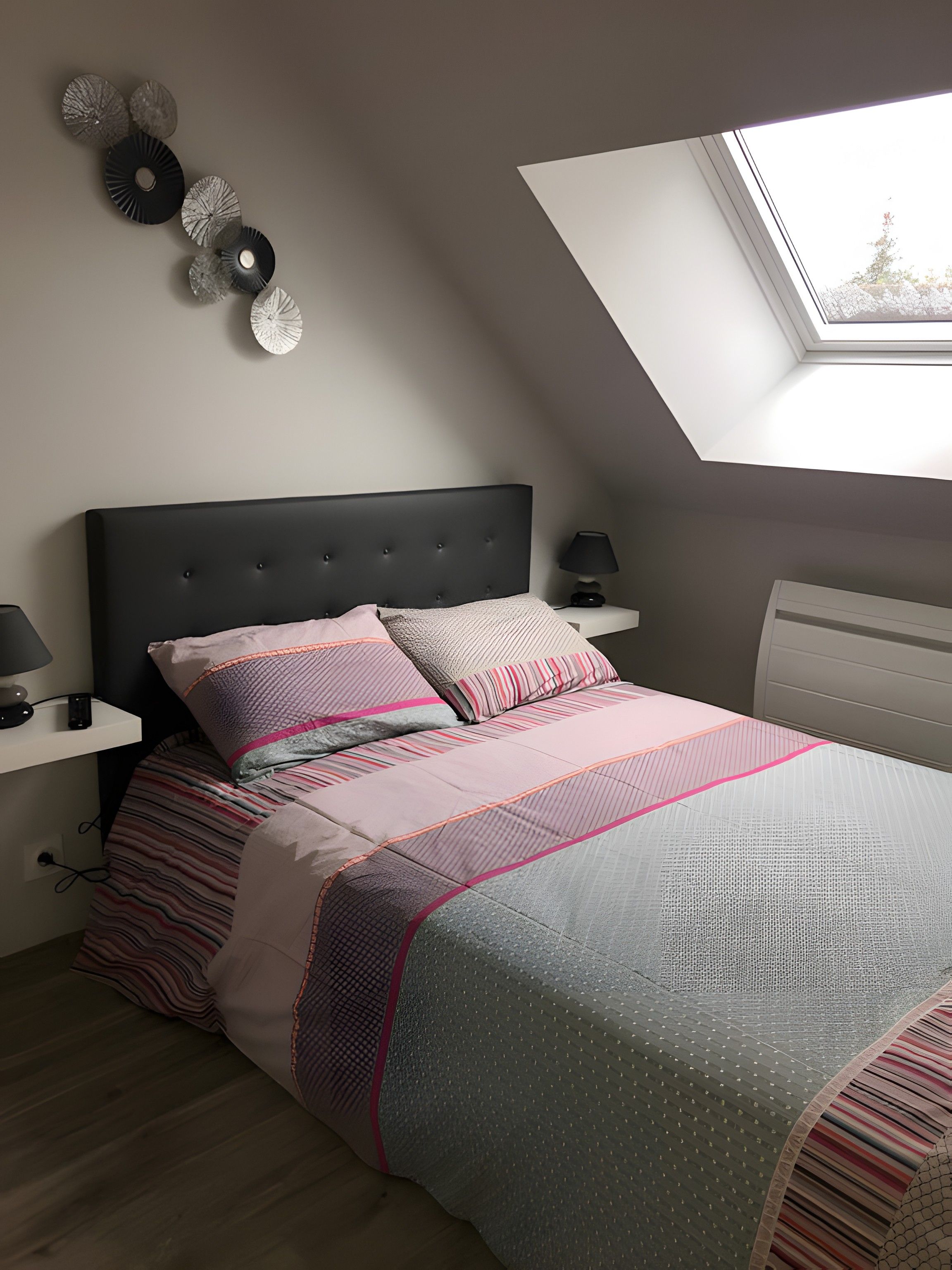 A bedroom with a sloped ceiling, a bed with patterned bedding, bedside lamps, and wall decor.